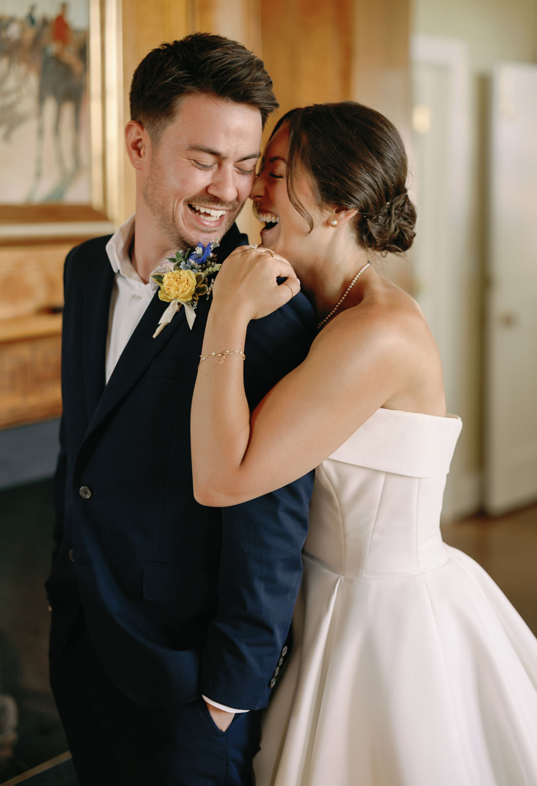 Bride and groom laughing together, her head resting on his shoulder in a candid indoor moment