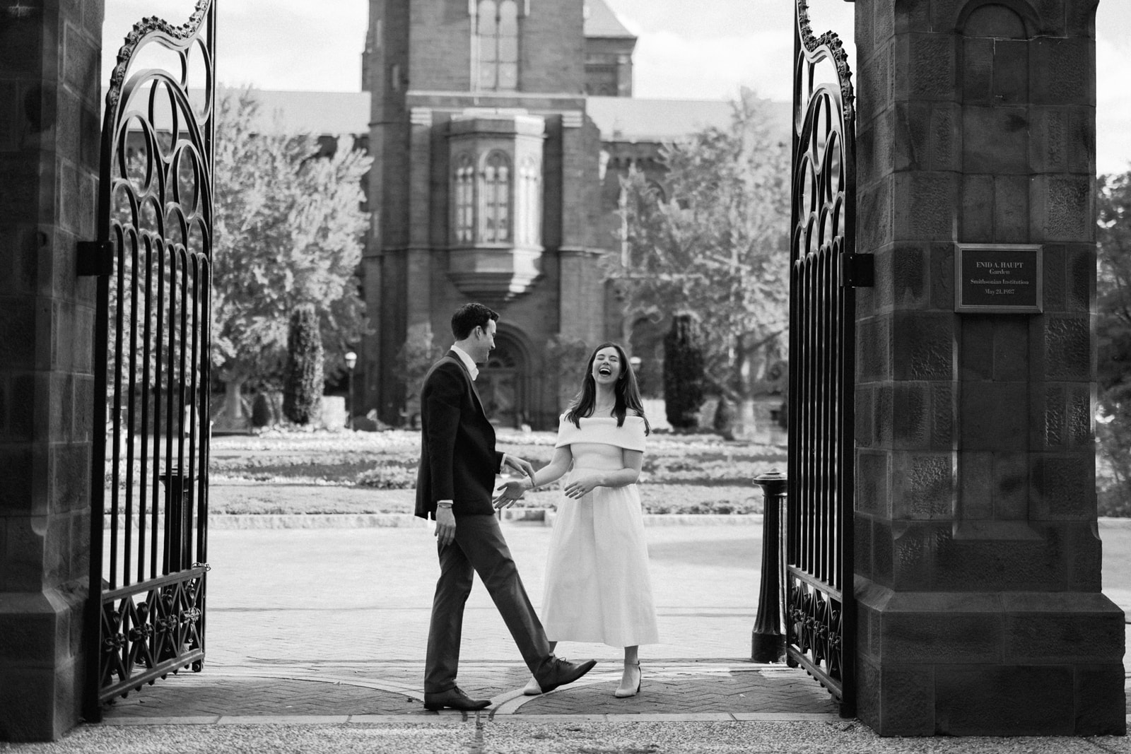 Black and white engagement photo of a couple walking past the iron gates of the Smithsonian Castle